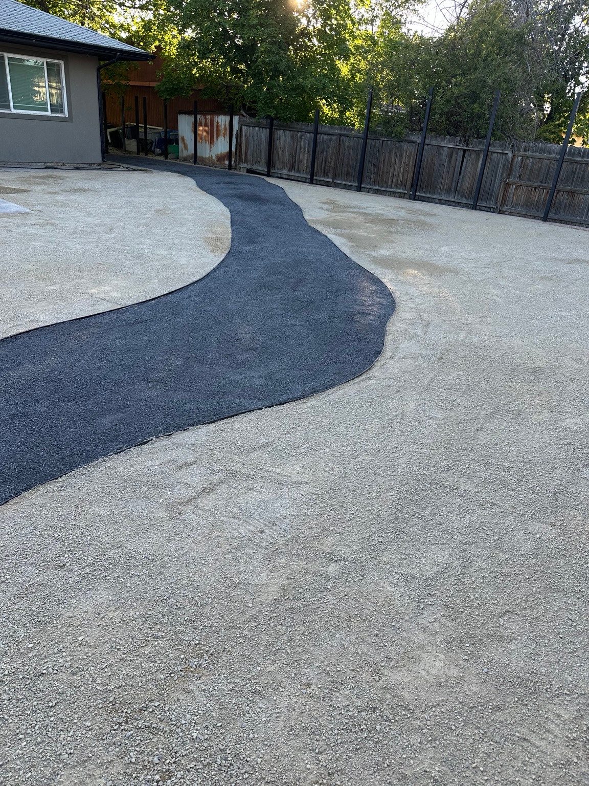 A dark curving path crosses a light-colored aggregate area beside a wooden fence.